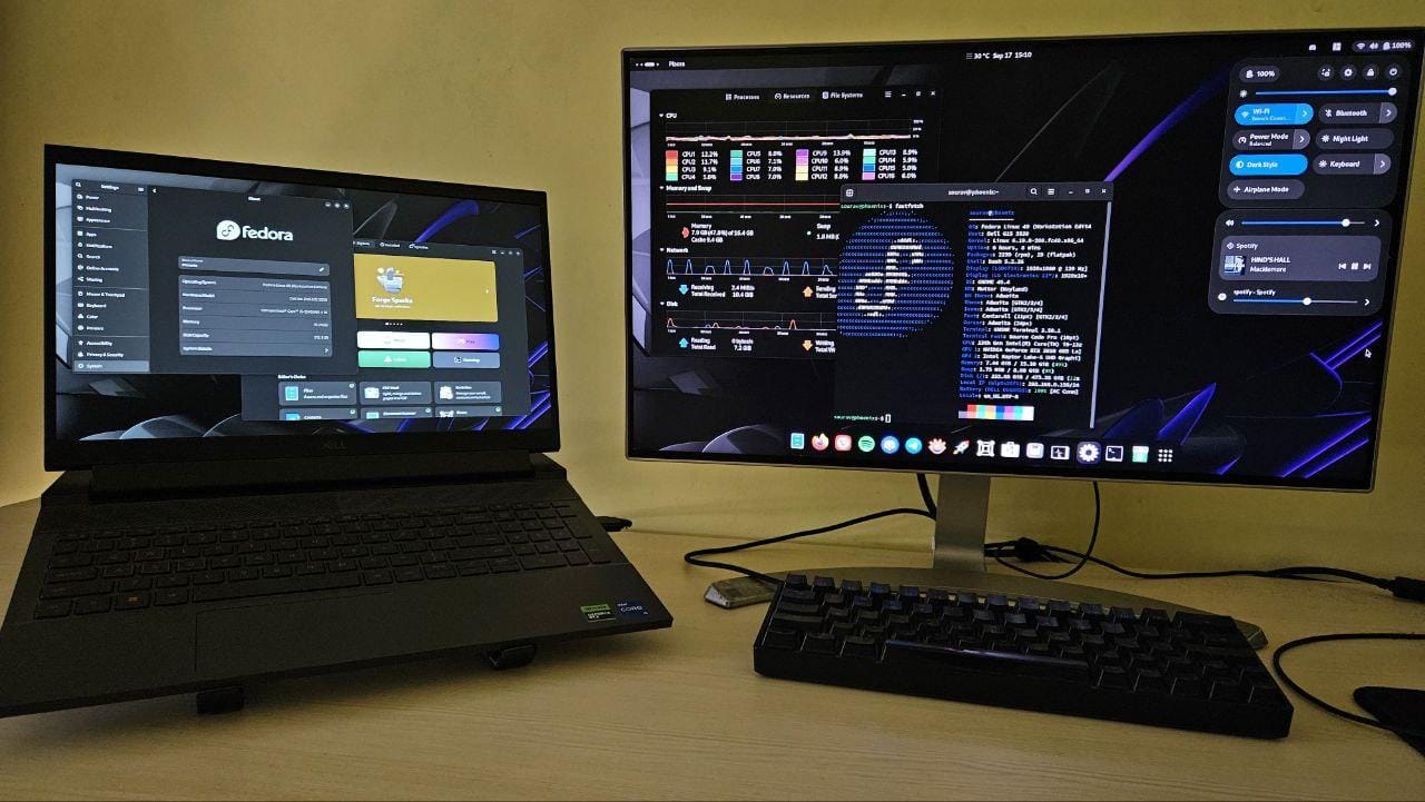 a photo showing a laptop with an external monitor running fedora 40 with some applications and a fastfetch output visible there is a keyboard and some wires in the photo too